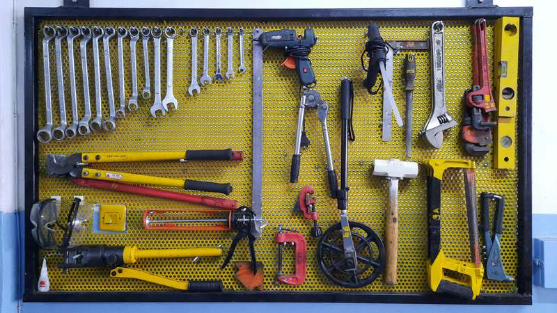 Side view of a person wearing safety glasses and gloves tightening a cabinet hinge with a cordless drill on a workshop bench